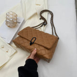 A tan quilted flap shoulder bag with a black metal clasp and a brown-and-black chain strap, held by a hand over a cream-colored blazer with a decorative glass vase and book in the background.