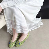 A person seated indoors wearing a long-sleeve white pleated midi skirt and light olive green flat shoes with blue and green rhinestone embellishments. A black display banner with Chinese text appears at the top, and a beige floor and dark chair are visible in the background.
