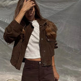 Woman wearing a brown jacket and pants with a white top, holding a woven bag against a plain background.
