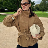 A woman wearing a tan suede-like belted jacket and black pants stands outdoors in a park, holding a cream-colored textured clutch bag.