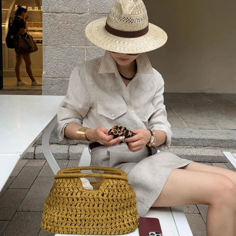 Woman sitting on a bench wearing a straw hat and holding a clutch, with a woven yellow bag beside her.