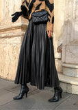 Person wearing a black pleated skirt, brown coat, and black boots on a stone pavement.