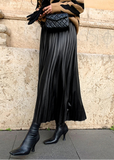 A person wearing a long black pleated skirt, black pointed-toe heeled boots, and a black quilted handbag, standing against a stone wall.